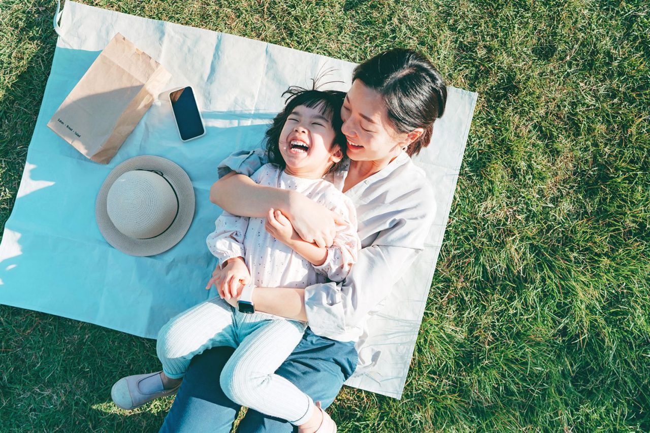 Affectionate young Asian mother embracing little daughter in arms, lying down on the grassy field, having fun and smiling joyfully, enjoying together on a sunny day. Family love and bonding time. Enjoying the nature. Outdoor fun concept