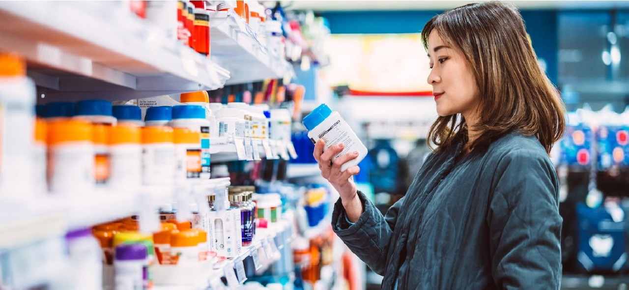 Beautiful Asian woman reading the ingredient label while shopping for multi vitamin & health supplements on shelf in supermarket. Heathy lifestyle concept.