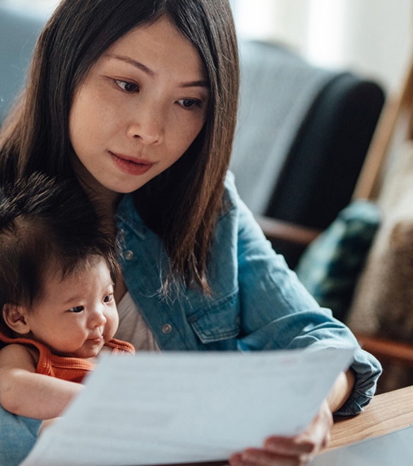Young Asian mother with baby daughter working on her laptop from home while holding her baby girl in her arm. Young working parent doing her home finances in the daytime at the living room.